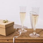 Indpakket gaveæske med guldsløjfe ved siden af to glas mousserende vin på et træbord