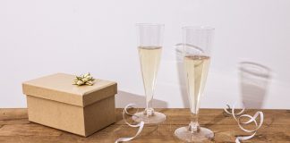 Indpakket gaveæske med guldsløjfe ved siden af to glas mousserende vin på et træbord
